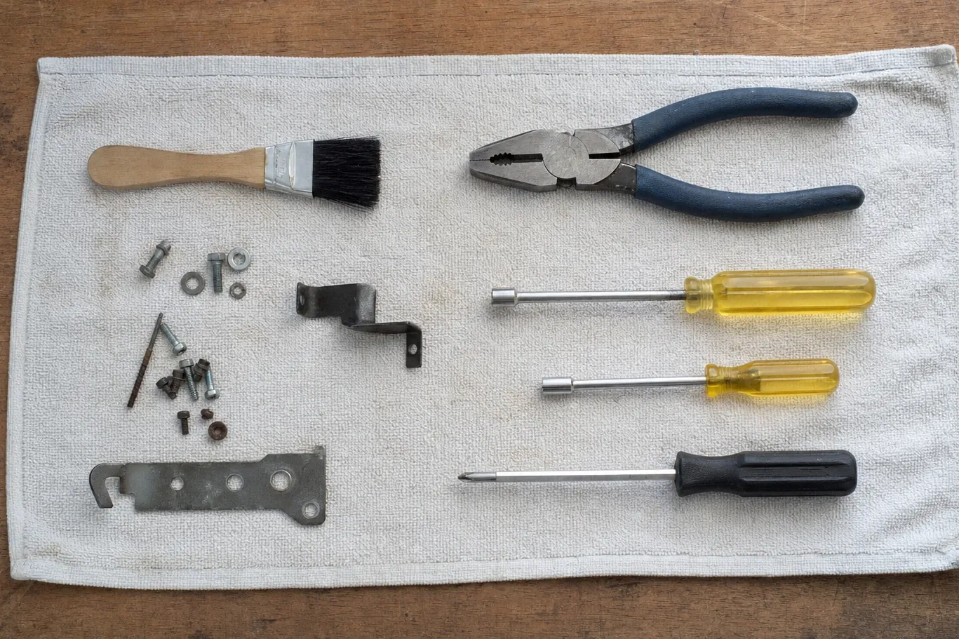 Bench flat-lay on a white towel: a bristle brush, pliers, two nut drivers, a screwdriver, a steel bracket, a drilled latch plate, bolts, washers and small screws