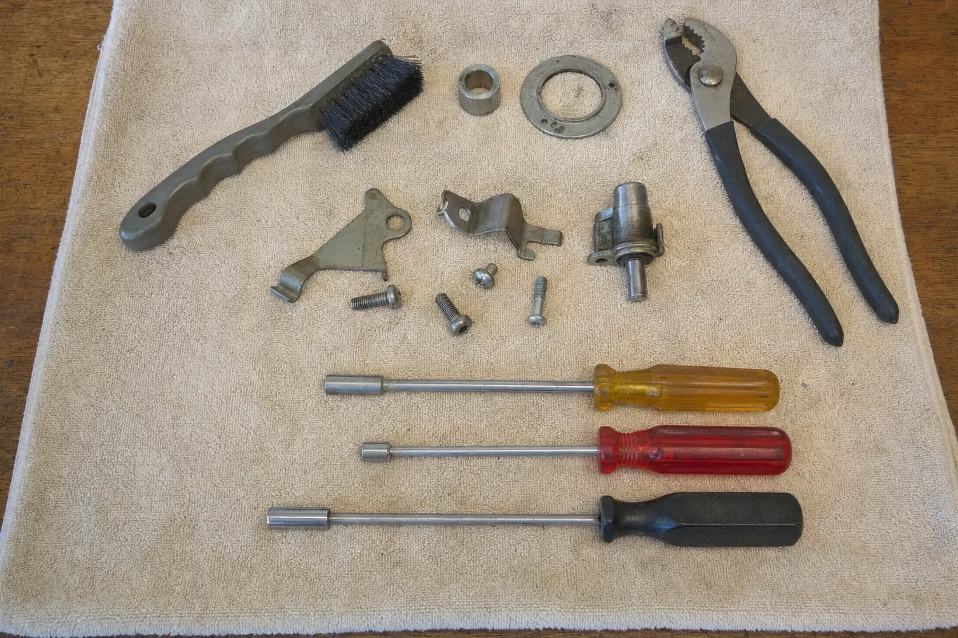 Bench flat-lay on a pale towel: a wire brush, a collar and a burner ring, slip-joint pliers, two brackets, a valve body with its shaft, machine screws and three nut drivers
