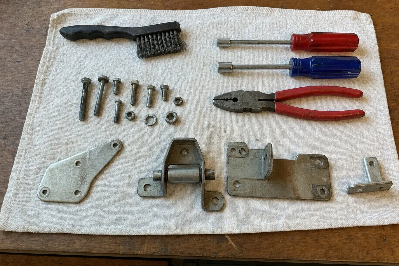 Bench flat-lay on a pale towel: a wire brush, bolts, nuts and washers, two nut drivers, red-handled pliers and four pieces of steel door hardware including a hinge with its roller